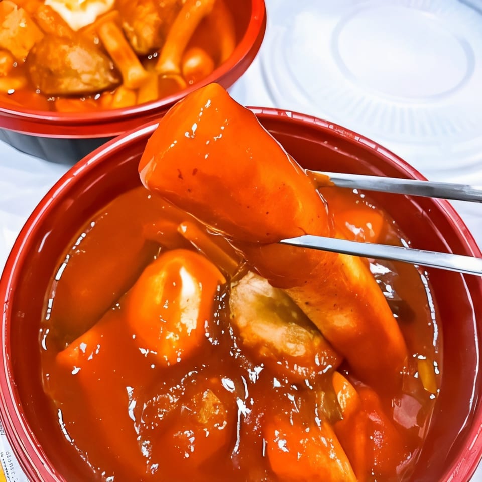 Chopsticks lifting a piece of tteokbokki from a red bowl of spicy Korean rice cakes.